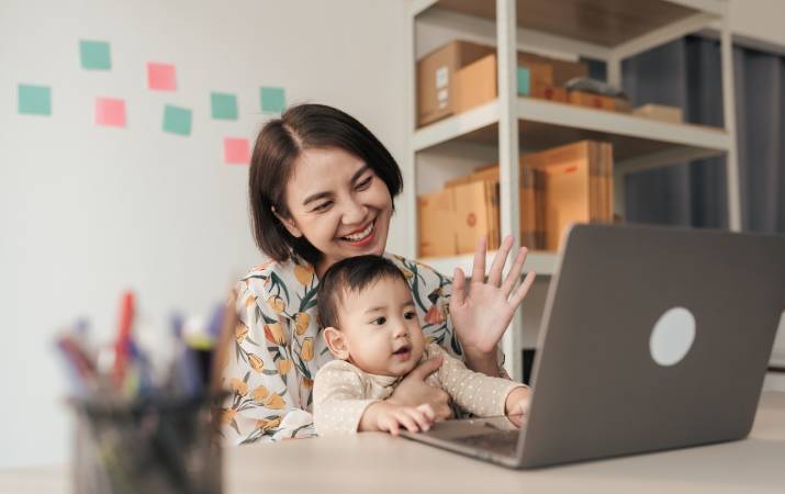 A happy tech professional greeting her colleagues over a laptop with her baby on her lap
